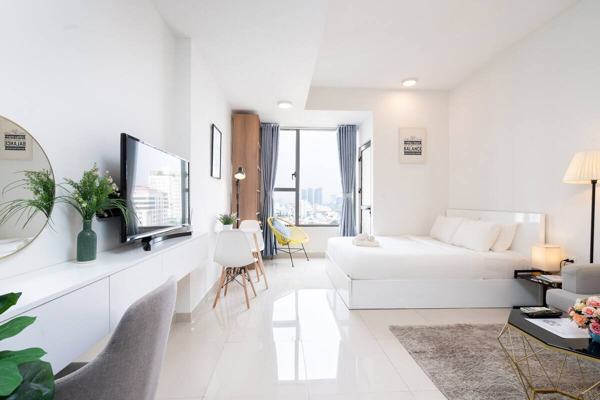 A modern hotel room with a large bed, a desk with a flat-screen TV, a dining area with chairs, and a view of the city through large windows.