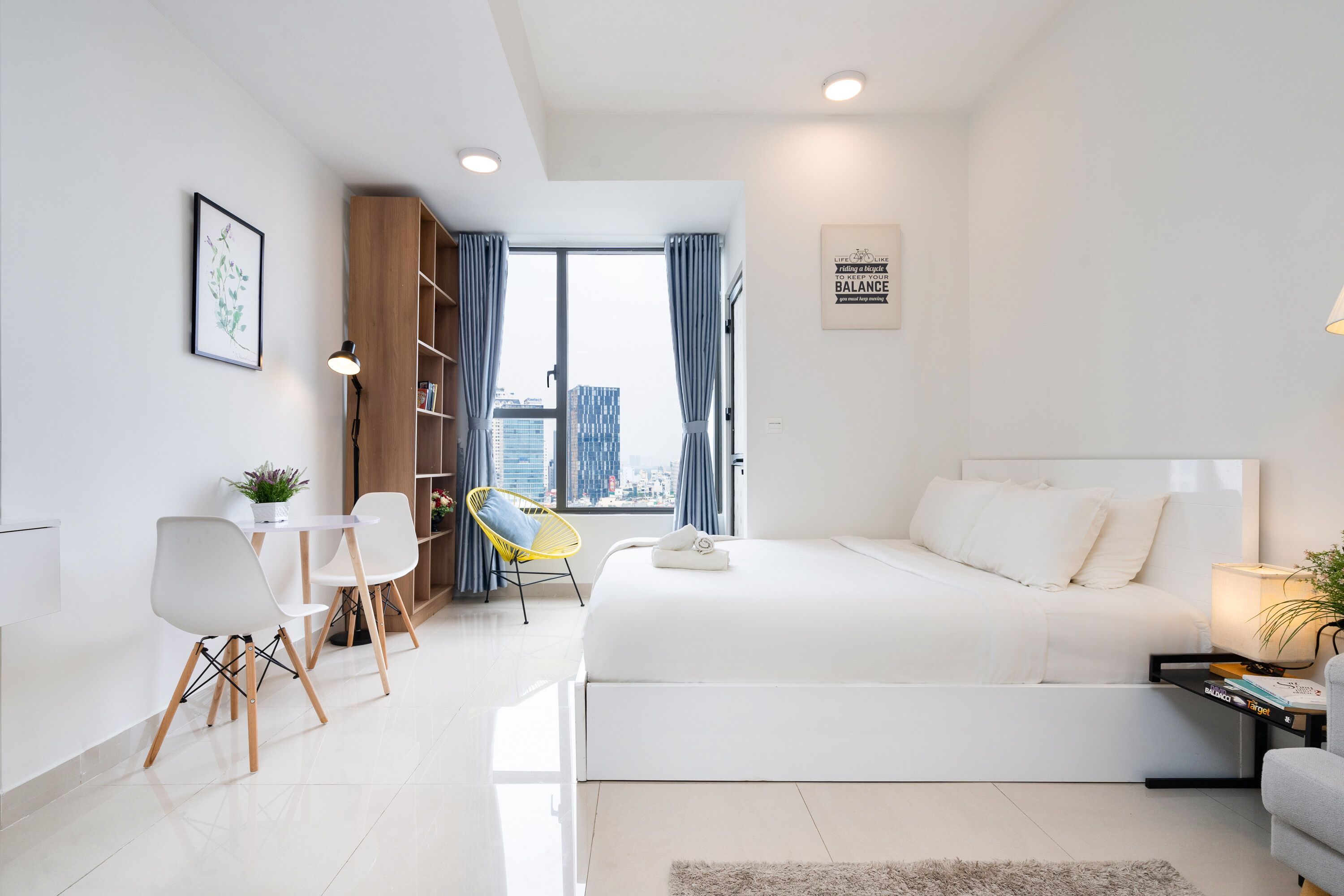 Une chambre moderne avec un grand lit, un bureau avec des chaises, une bibliothèque et une vue sur la ville par la fenêtre.