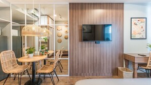 A modern hotel room with a wooden floor, a bed, a TV mounted on the wall, wicker chairs, a dining table, and a kitchen area visible through a glass partition. - TeeUP Home (Ho Chi Minh City)