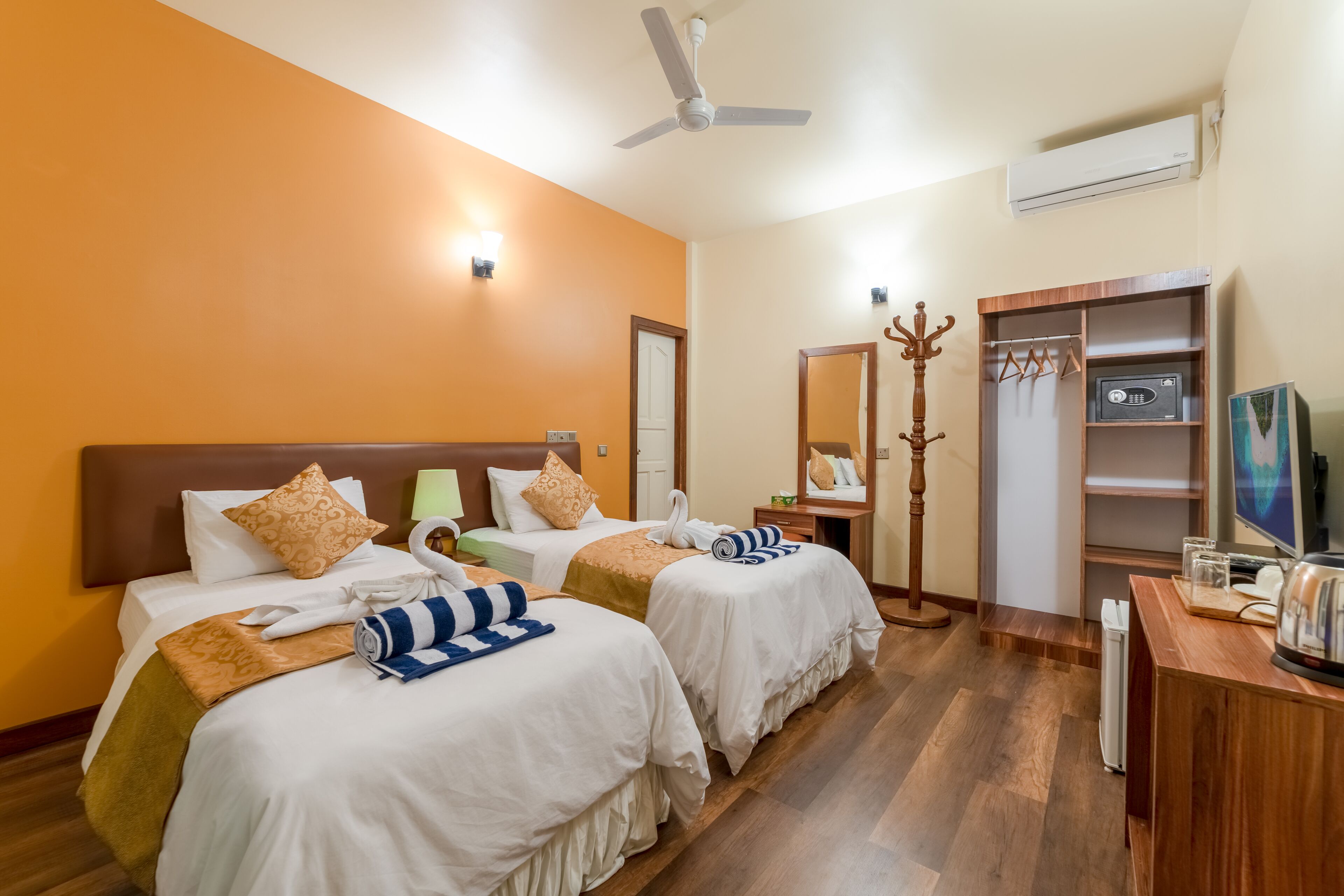 A hotel room with two beds, a ceiling fan, an air conditioning unit, a wooden coat rack, a television, and a desk with a computer.