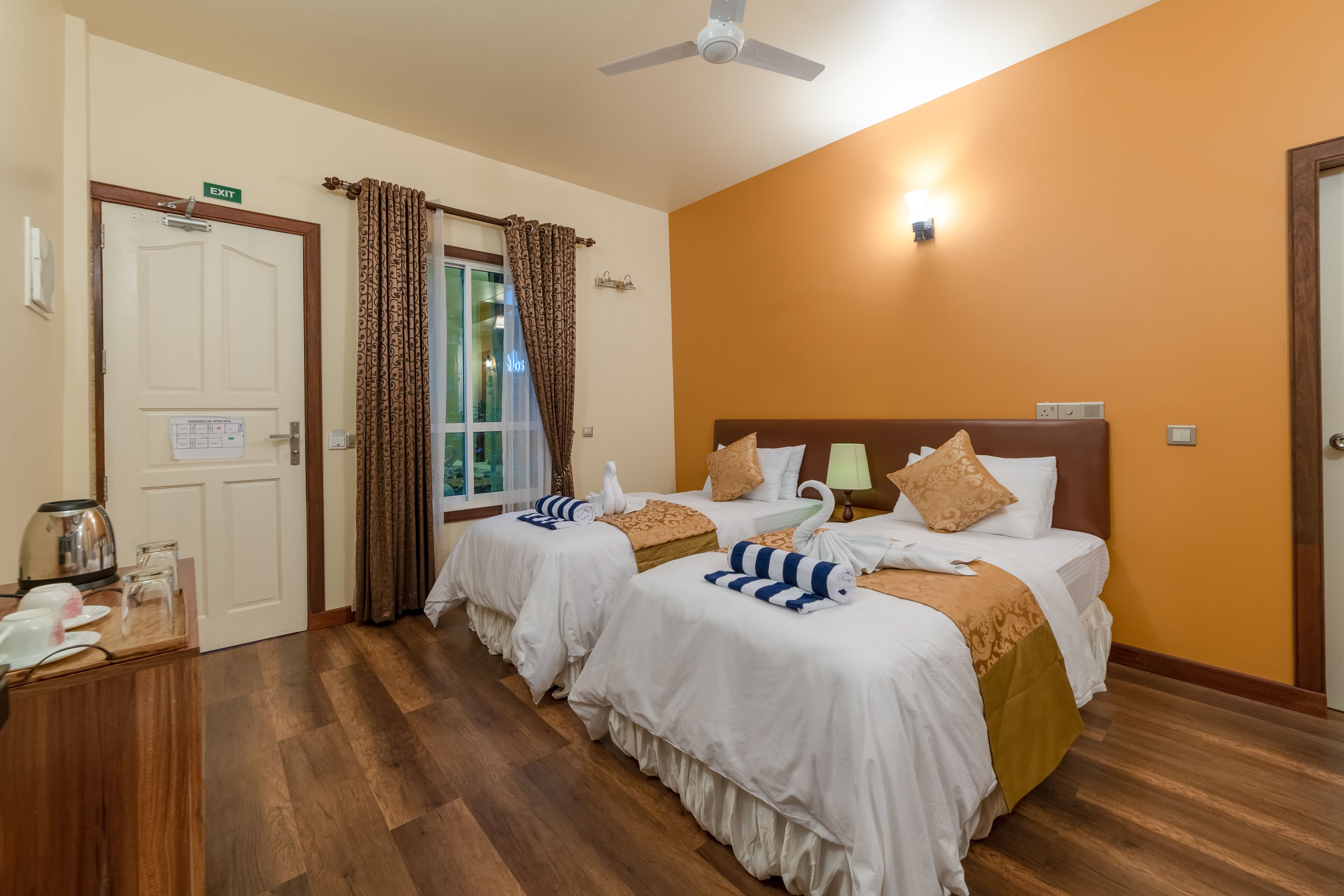Habitación de hotel con dos camas, una ventana con cortinas, un ventilador de techo y una pequeña mesa con una tetera y platos.