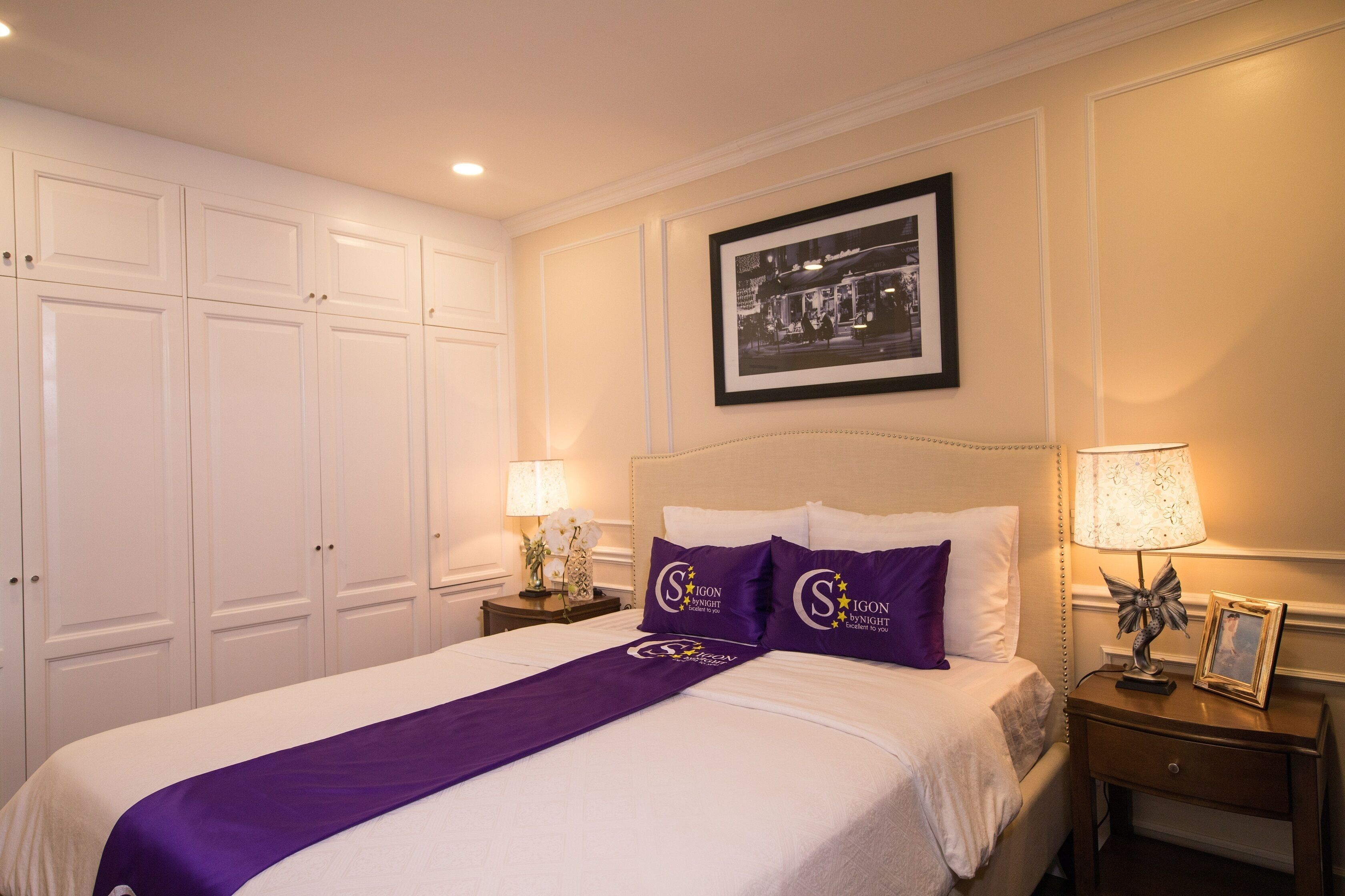 A neatly made bed with a purple bedspread and matching pillows, a framed picture above the bed, and a nightstand with a lamp and a framed photo.