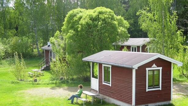 A small cabin with a porch, surrounded by lush greenery. - Falkudden Camping (By Kyrkby)