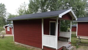 A small, single-story cabin with a porch and a door, surrounded by grass and trees. - Falkudden Camping (By Kyrkby)