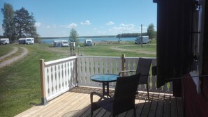 Une terrasse en bois avec une table et des chaises, donnant sur une zone herbeuse avec un lac et des véhicules garés.
