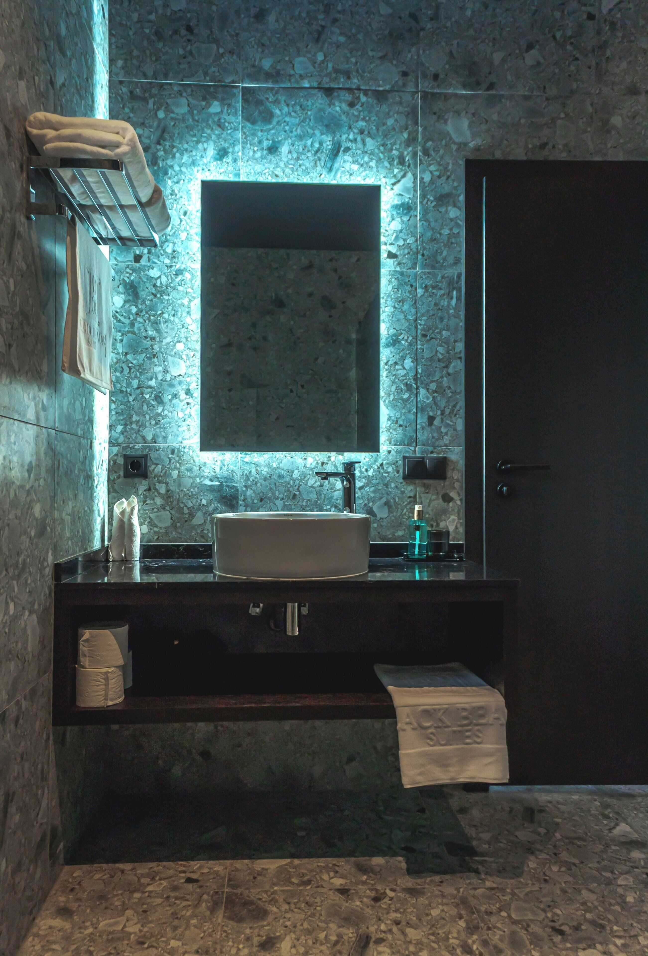A bathroom with a marble wall, a large mirror, a sink, and a towel rack.