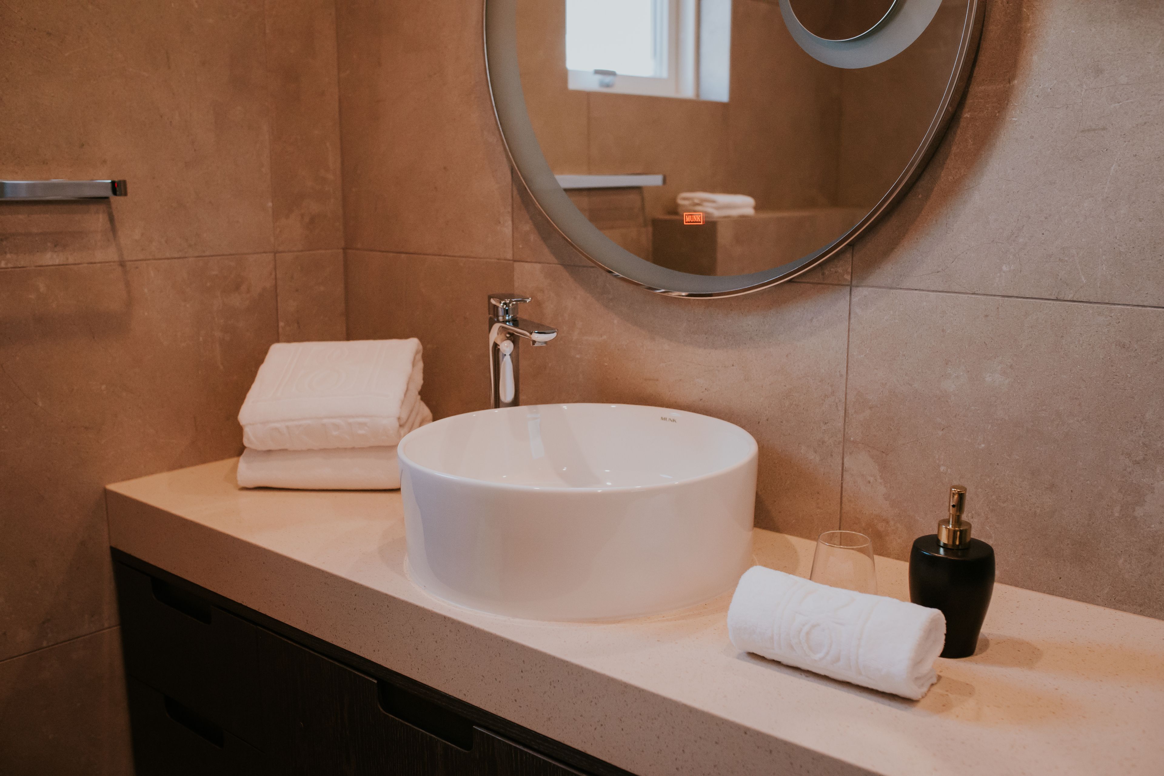 A bathroom with a round white sink, a mirror, a towel, and a bottle of soap.