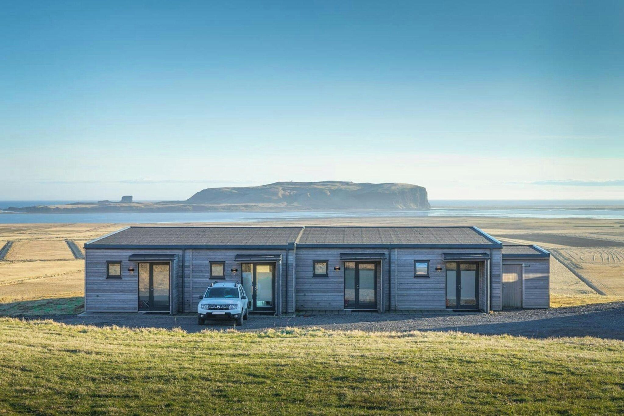 A modern two-story building with a car parked in front, situated on a grassy field with a view of a distant hill and water body.