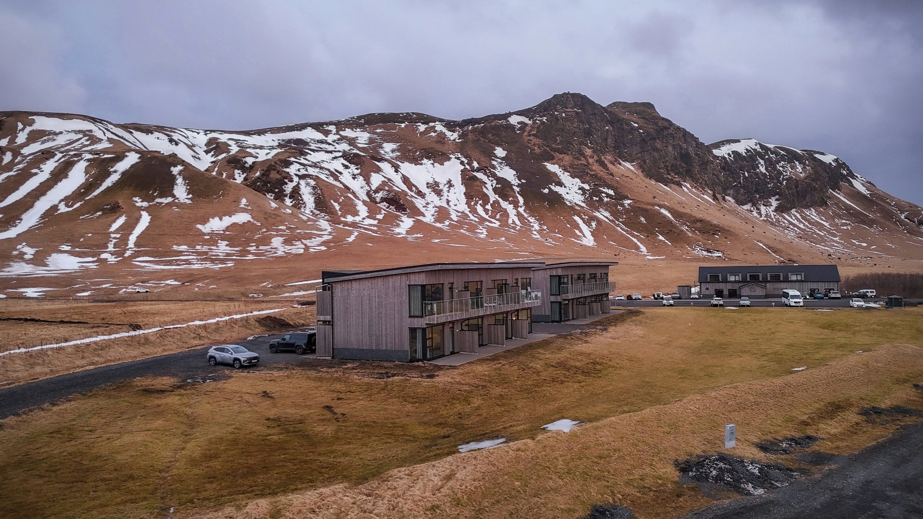 A modern building with a flat roof and large windows, situated in a grassy area with a mountain in the background.