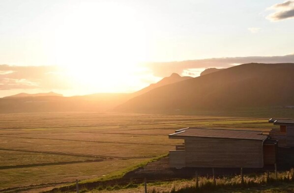 A modern building with a flat roof, situated in a rural landscape with expansive fields and distant mountains. - Black Beach Suites (Vik I Myrdal)