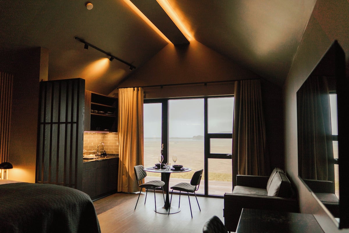 A room with a bed, a dining table with chairs, a sofa, a kitchen area, and large windows offering a view.