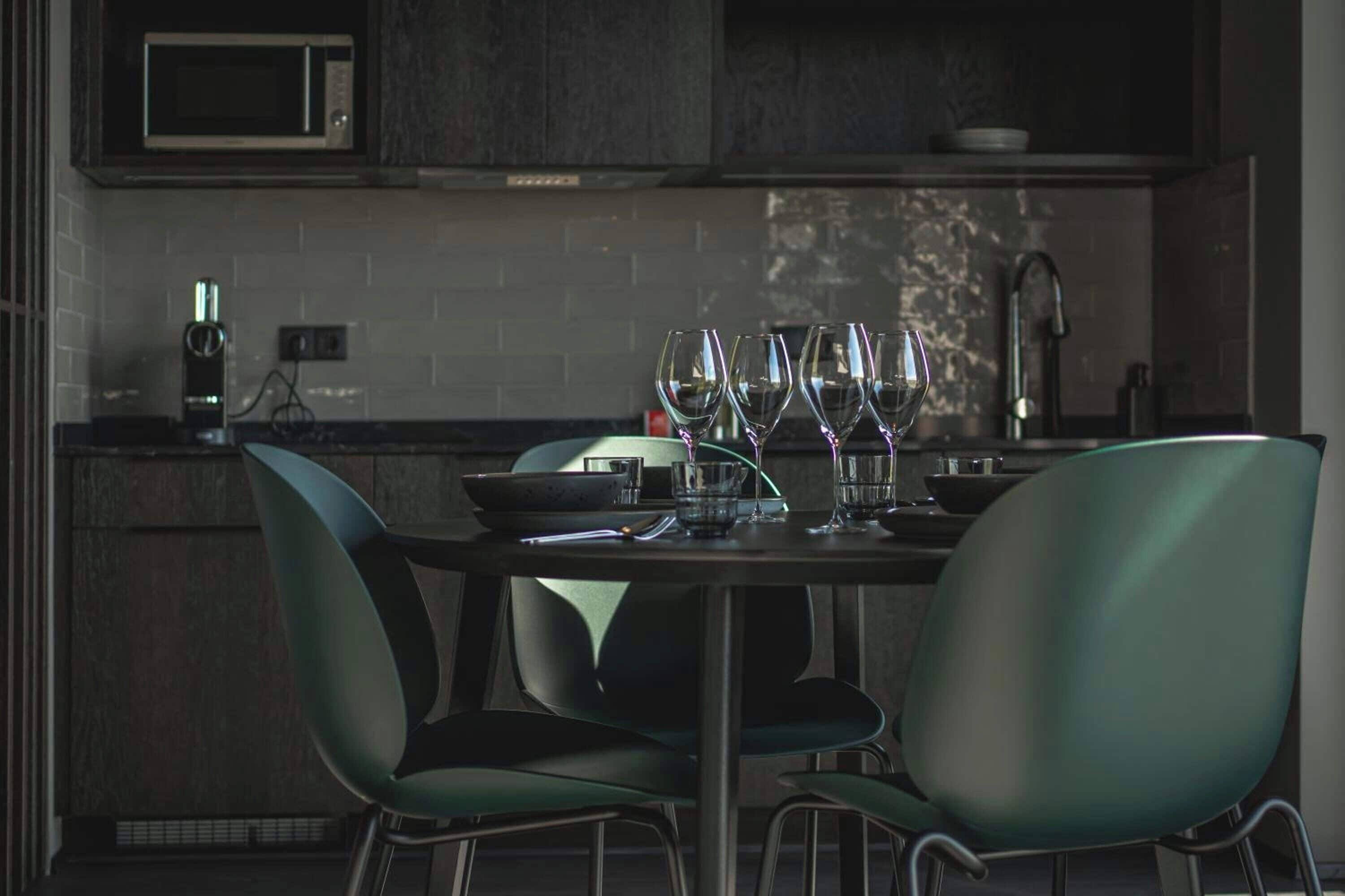 A modern kitchen with a dining table set for four, featuring green chairs and a central island with a bowl and glasses.