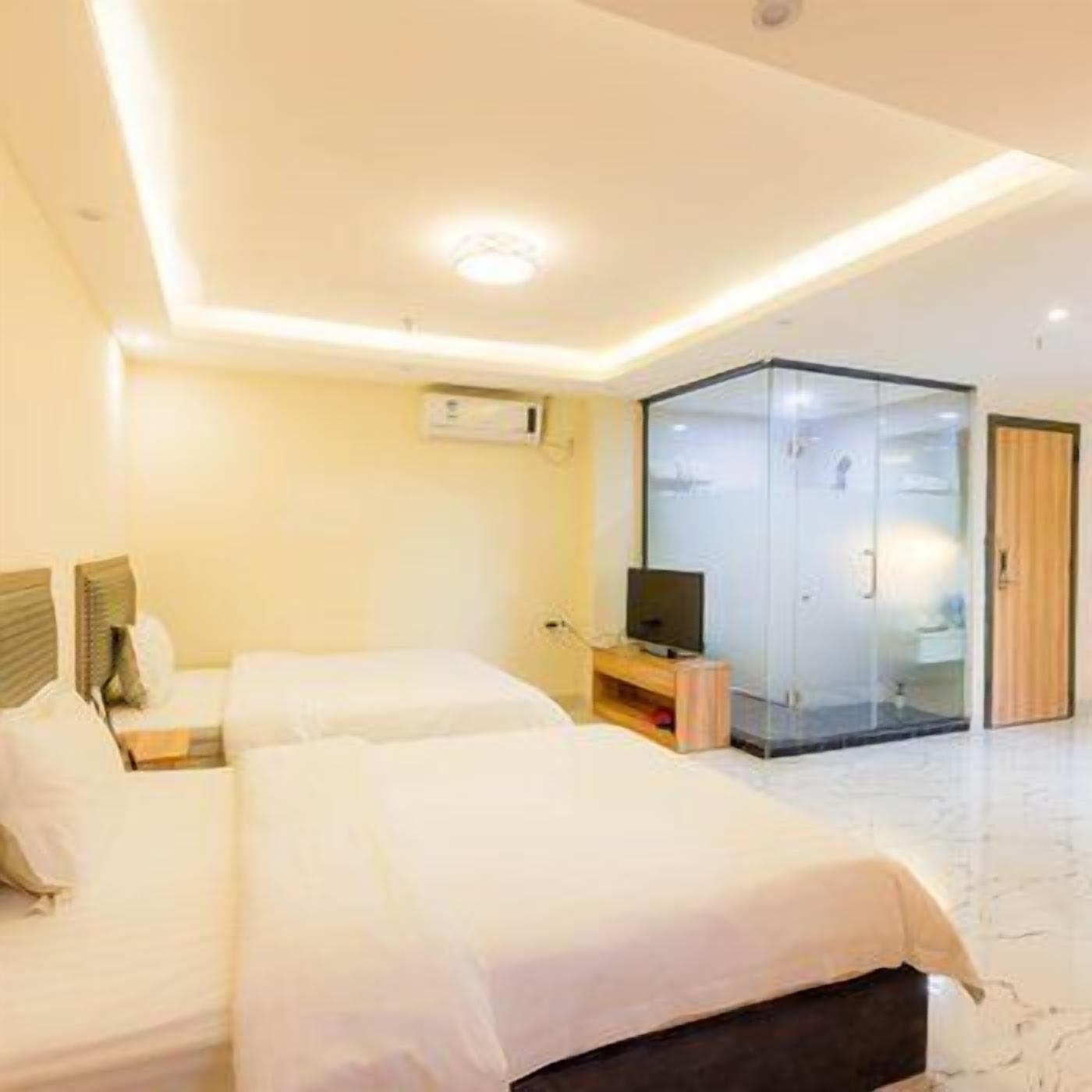A hotel room with two beds, a TV, a glass-enclosed bathroom, and a wooden door.