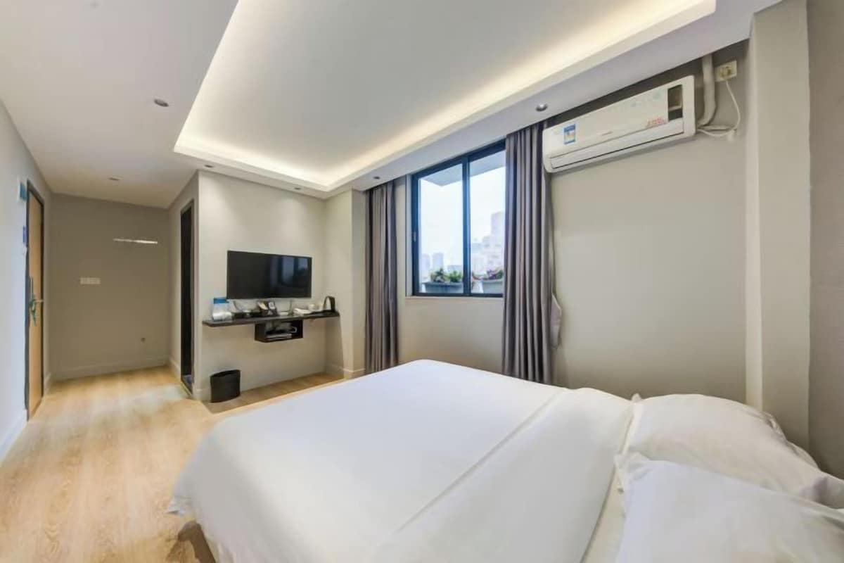 A hotel room with a bed, a TV, a window with curtains, and an air conditioning unit.