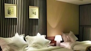 Habitación de hotel con dos camas, una colcha grande, una mesita de noche con lámpara y dos cuadros en la pared.