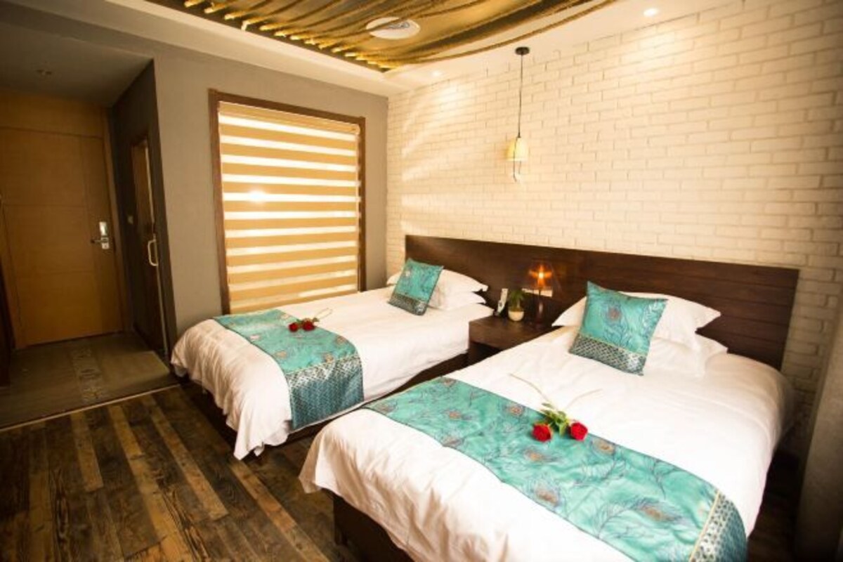 A hotel room with two beds, wooden flooring, a brick wall, and a window with blinds.