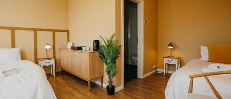 Habitación de hotel con dos camas, un aparador de madera, una planta en maceta y la puerta del baño.