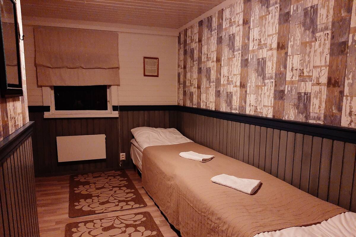Ein schmaler Raum mit einem Einzelbett, einem Fenster mit Jalousie, einem Heizkörper und einer Wand mit Muster.