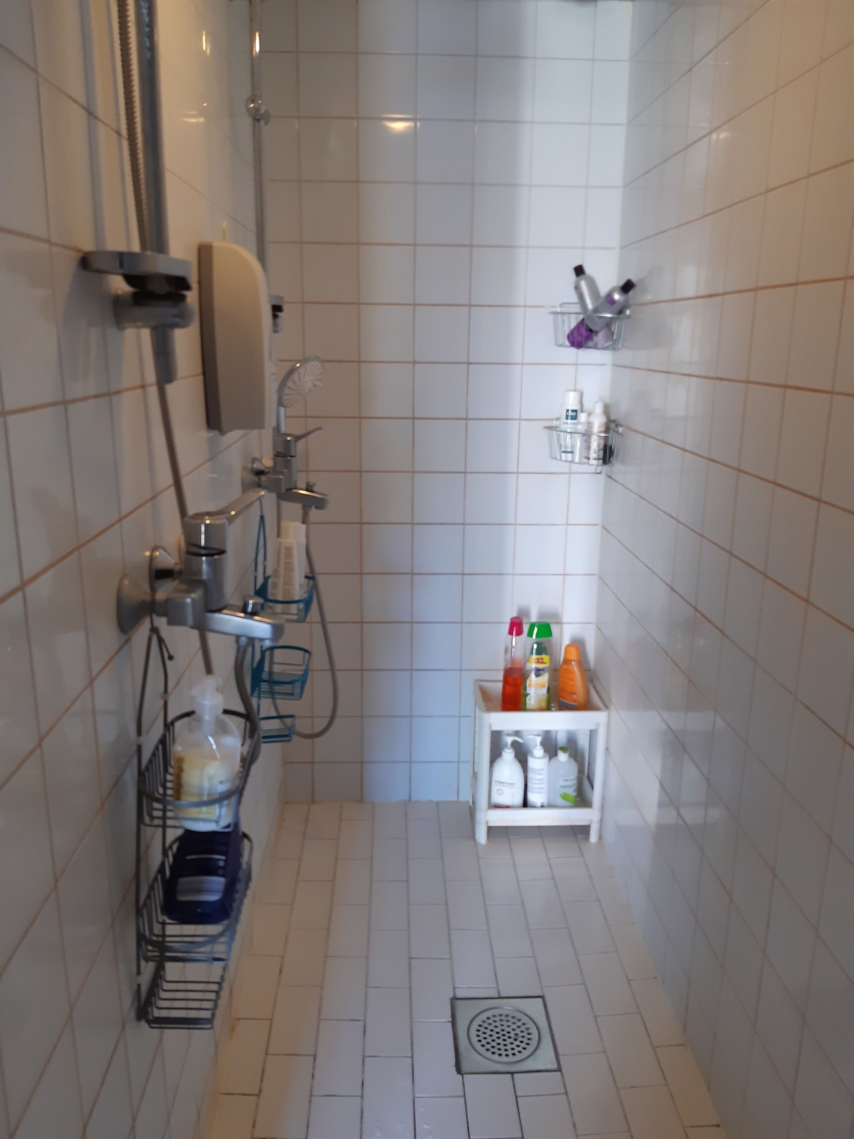 Un pequeño espacio de ducha con azulejos, un cabezal de ducha manual, un grifo empotrado en la pared y un estante con artículos de aseo.