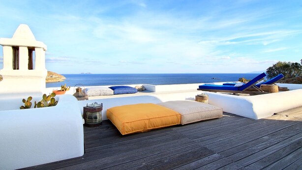 Una terrazza panoramica con vista libera sul mare, caratterizzata da un pavimento in legno, pareti bianche e poltrone da lounge blu.