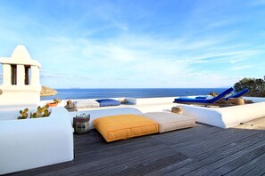 Um lounge no terraço com vista desobstruída para o mar, apresentando um deck de madeira, paredes brancas e confortáveis cadeiras de estar azuis.