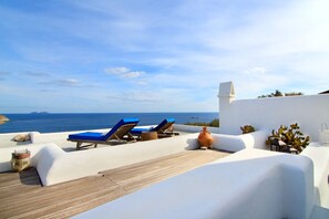 A rooftop terrace with white walls, blue lounge chairs, and a view of the sea.