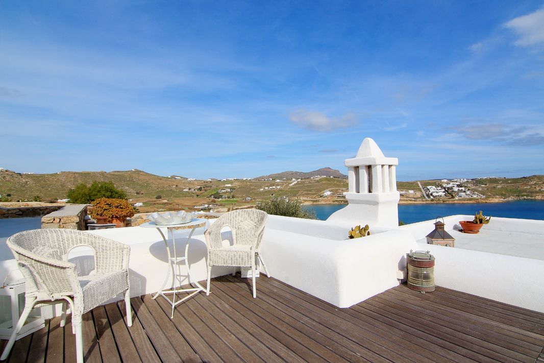 A terrace with white wicker chairs and a table, overlooking a coastal landscape with hills and a body of water.
