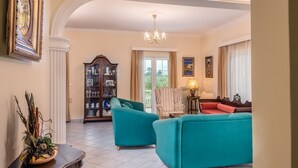 A spacious living room with a large window, a chandelier, and a classic wooden cabinet. - Zante 5 bedroom Villa with private pool and basketball court (Zakynthos)
