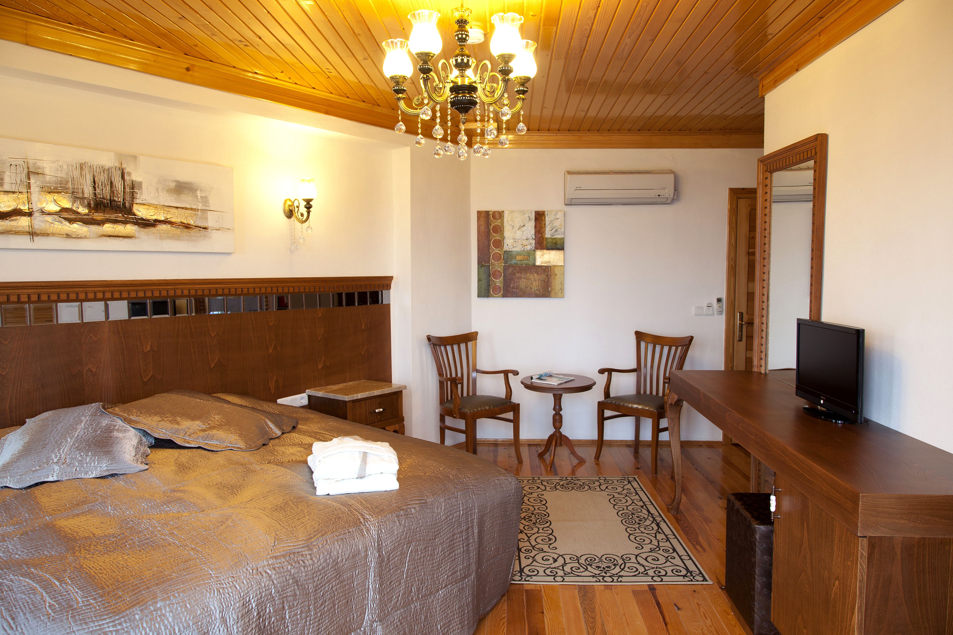 Una habitación de hotel con una cama grande, un cabecero de madera, una lámpara de araña, un televisor y una pequeña mesa con sillas.