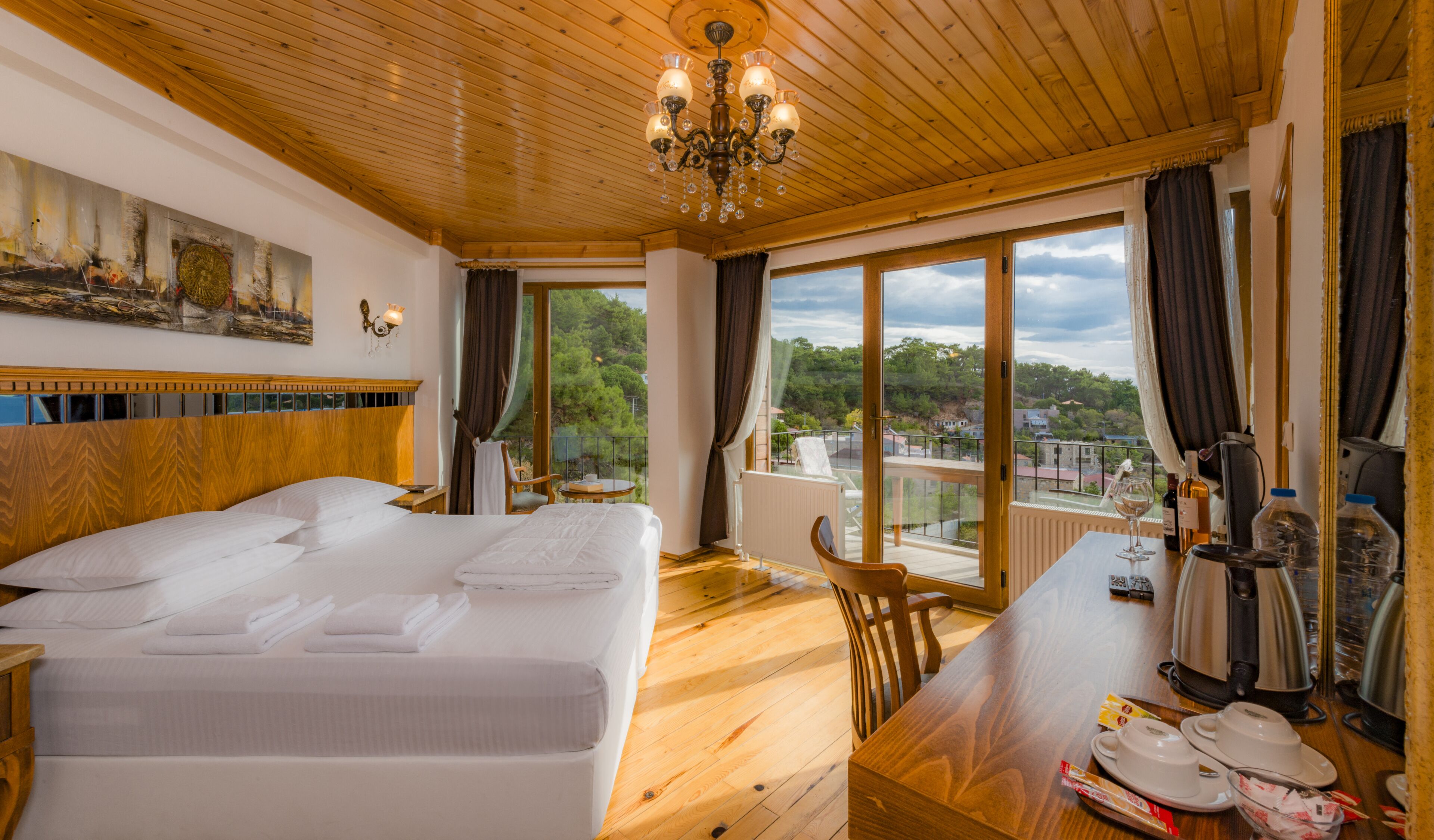 Habitación de hotel con una cama grande, techo de madera, candelabro, mesa de comedor con sillas y vistas al exterior.