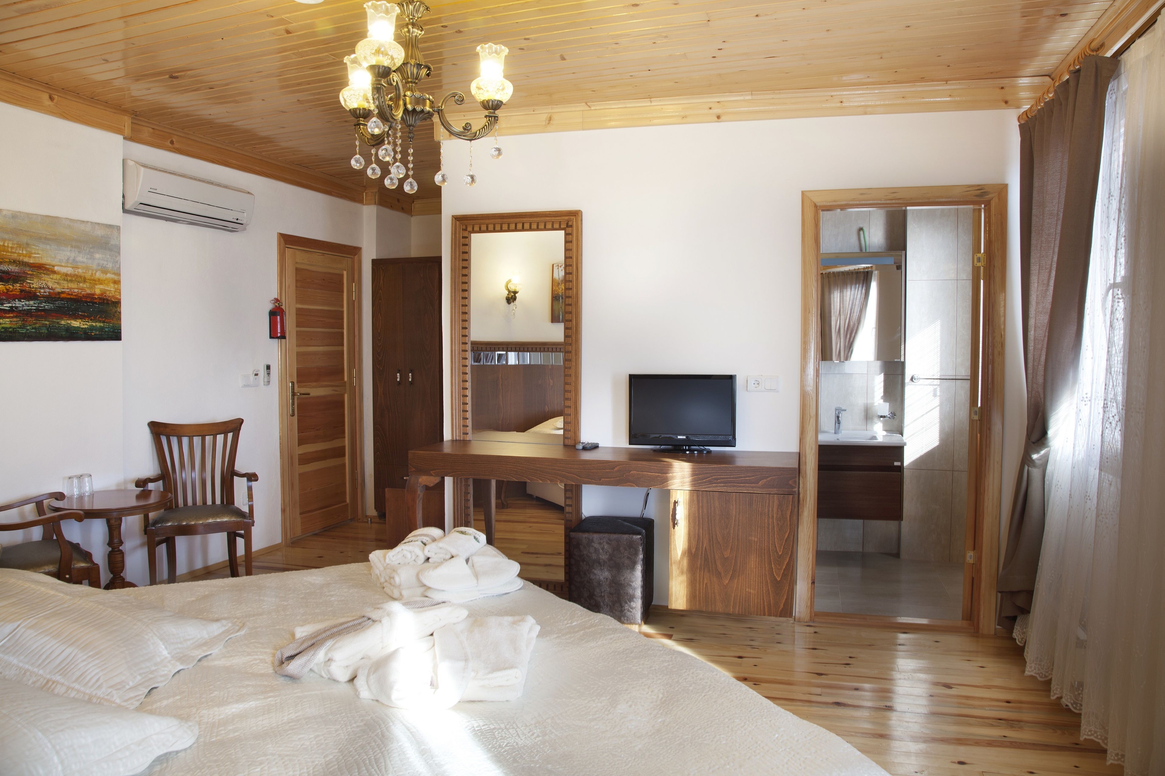 A hotel room with a wooden ceiling, a bed with white linens, a desk with a television, a chair, and a small table with a glass.