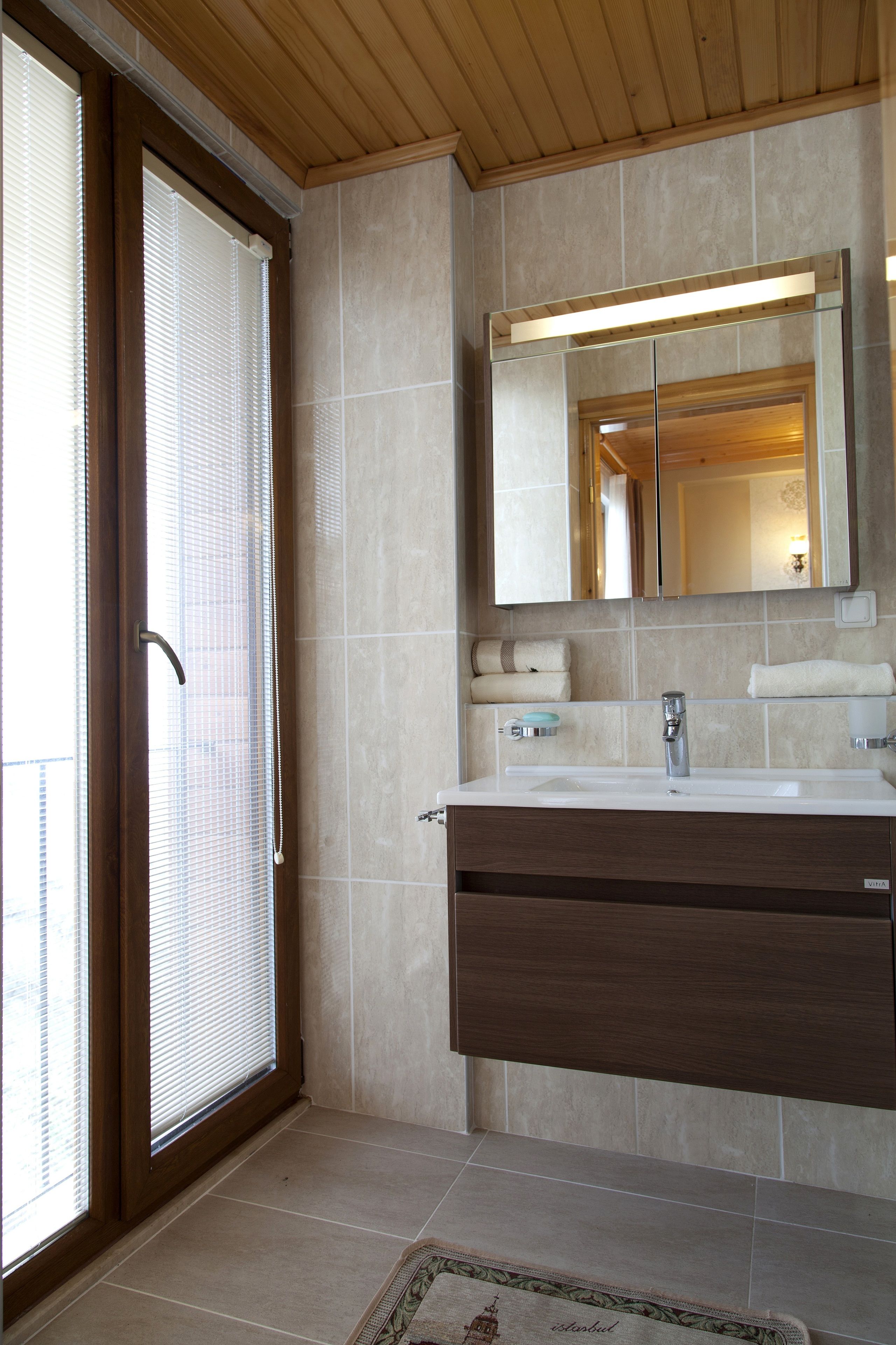 Ein Badezimmer mit Holzboden, einem großen Spiegel, einem Waschbecken mit Handtuchhalter und einem Fenster mit Jalousien.
