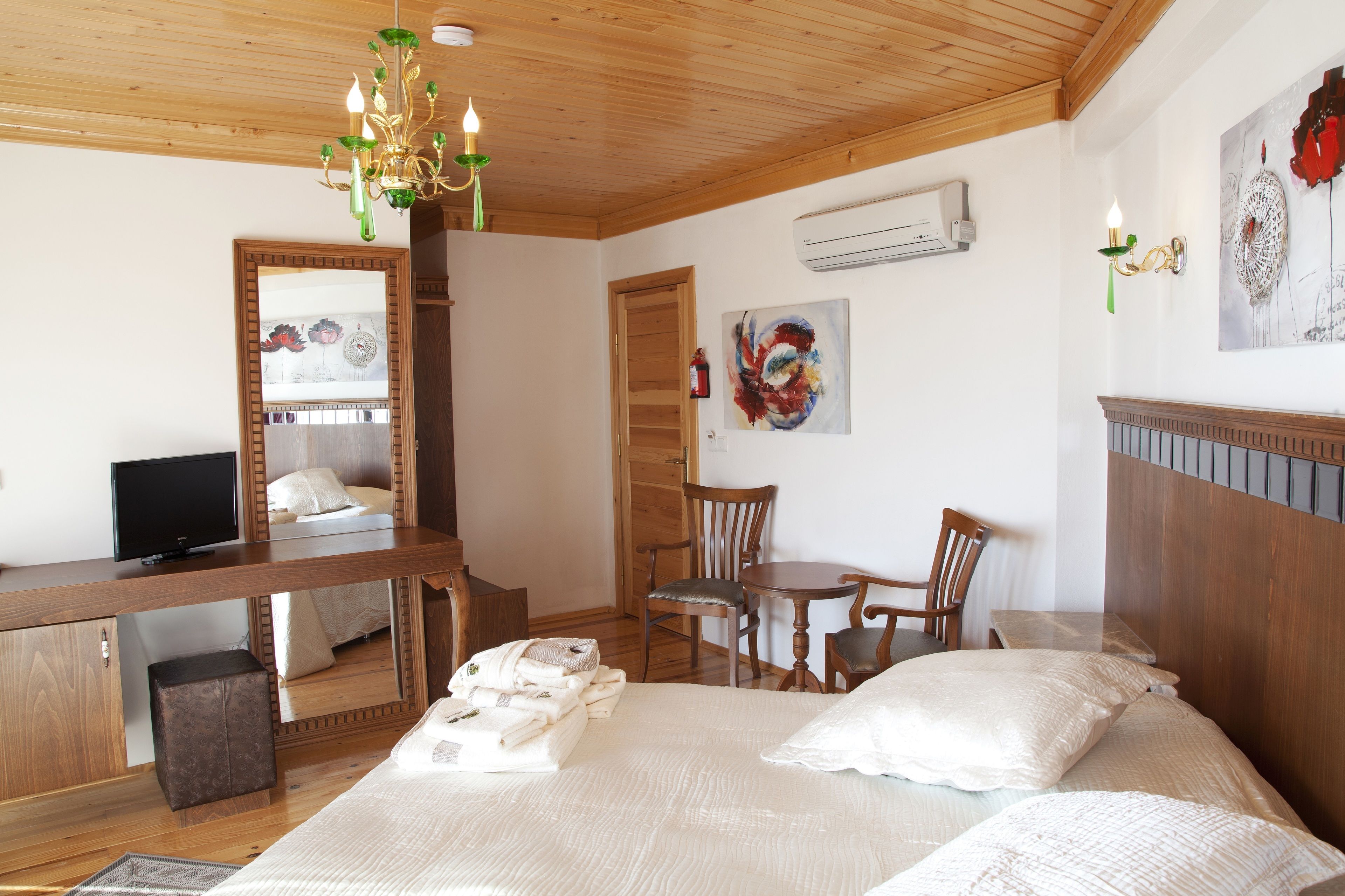 A bedroom with a wooden ceiling, a bed with white linens, a desk with a television, a chair, and a small table.