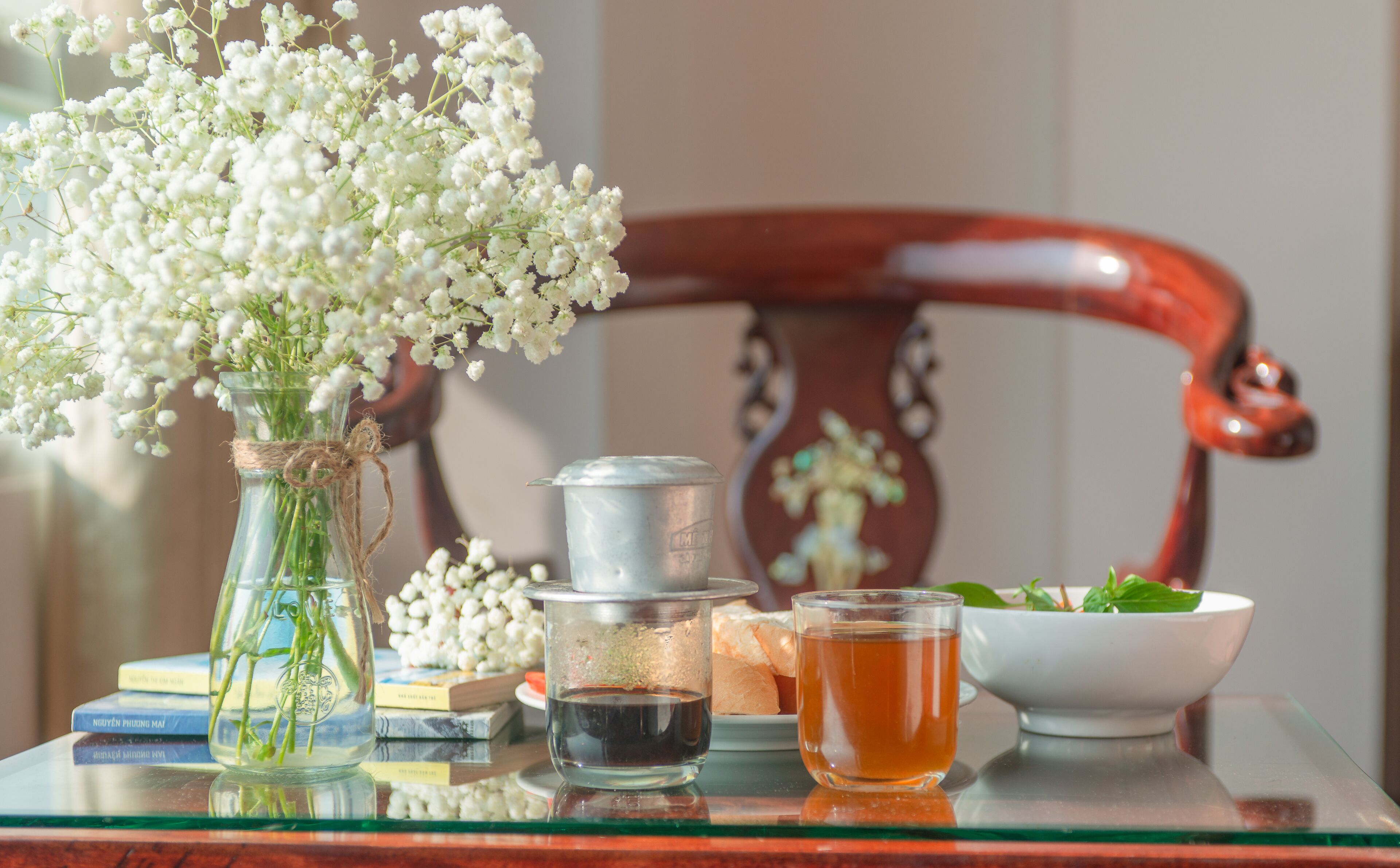 A glass vase with white flowers, a metal container, a glass with tea, and a bowl of greens on a reflective surface.