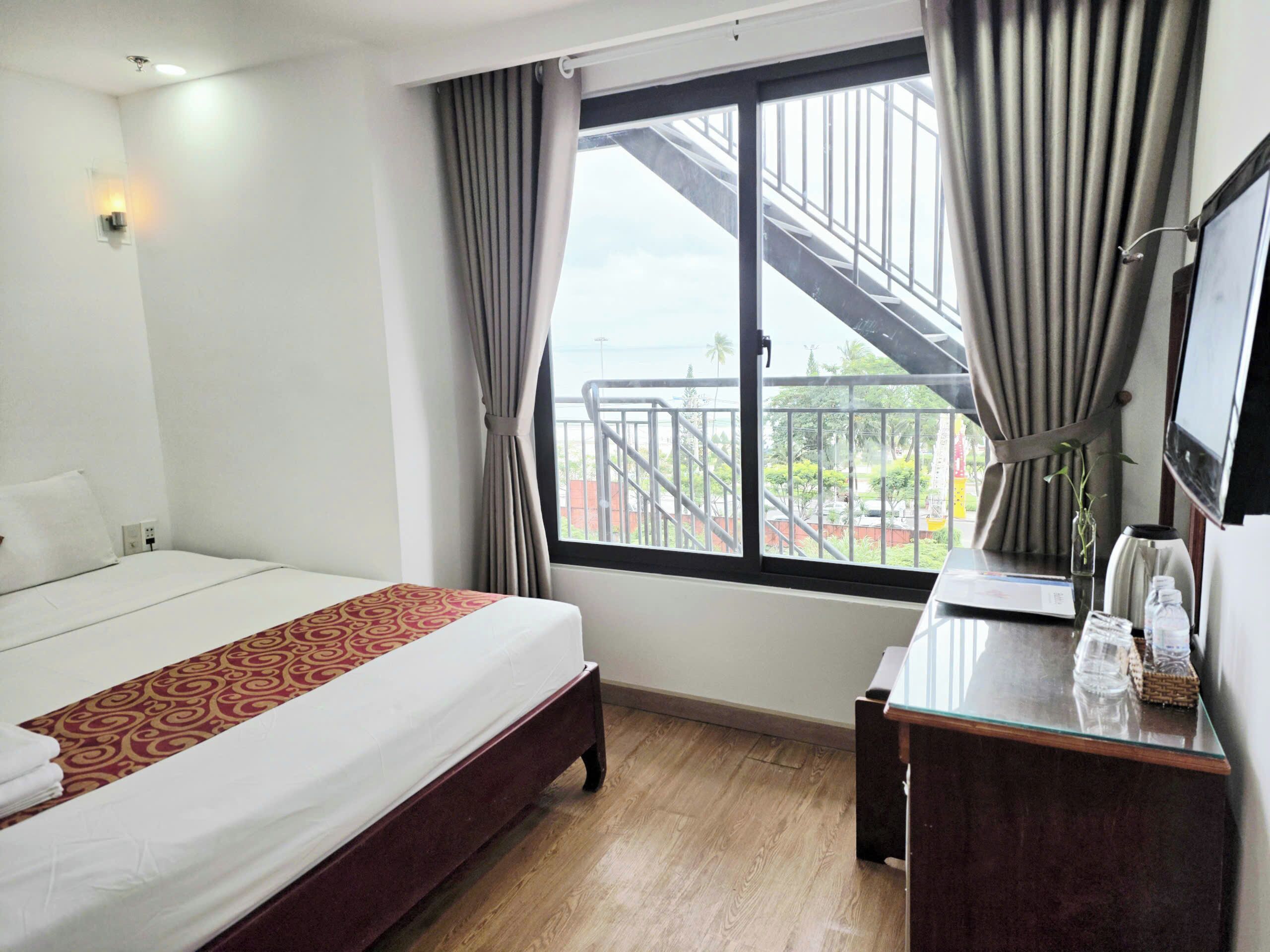 A hotel room with a bed, a desk, a mirror, and a window with a view of a balcony and outside scenery.