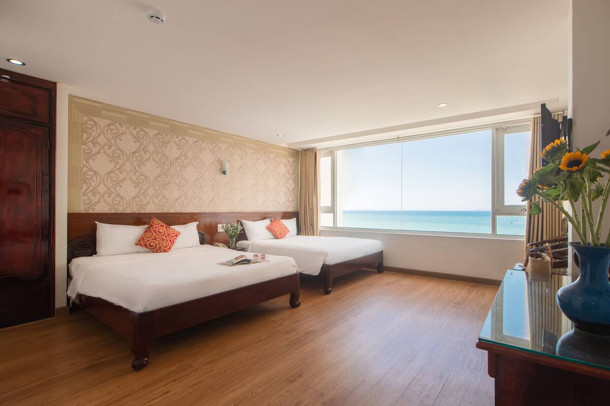 Une chambre d’hôtel avec deux lits, un plancher en bois, une grande fenêtre donnant sur la mer et un vase rempli de fleurs posé sur une table.