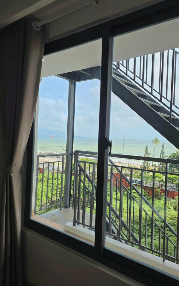A room with a large window overlooking a balcony with a view of a beach and palm trees.
