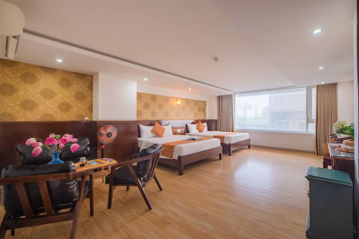 Een hotelkamer met twee bedden, een houten vloer, een bureau met stoel, een vaas met bloemen en een groot raam met gordijnen.