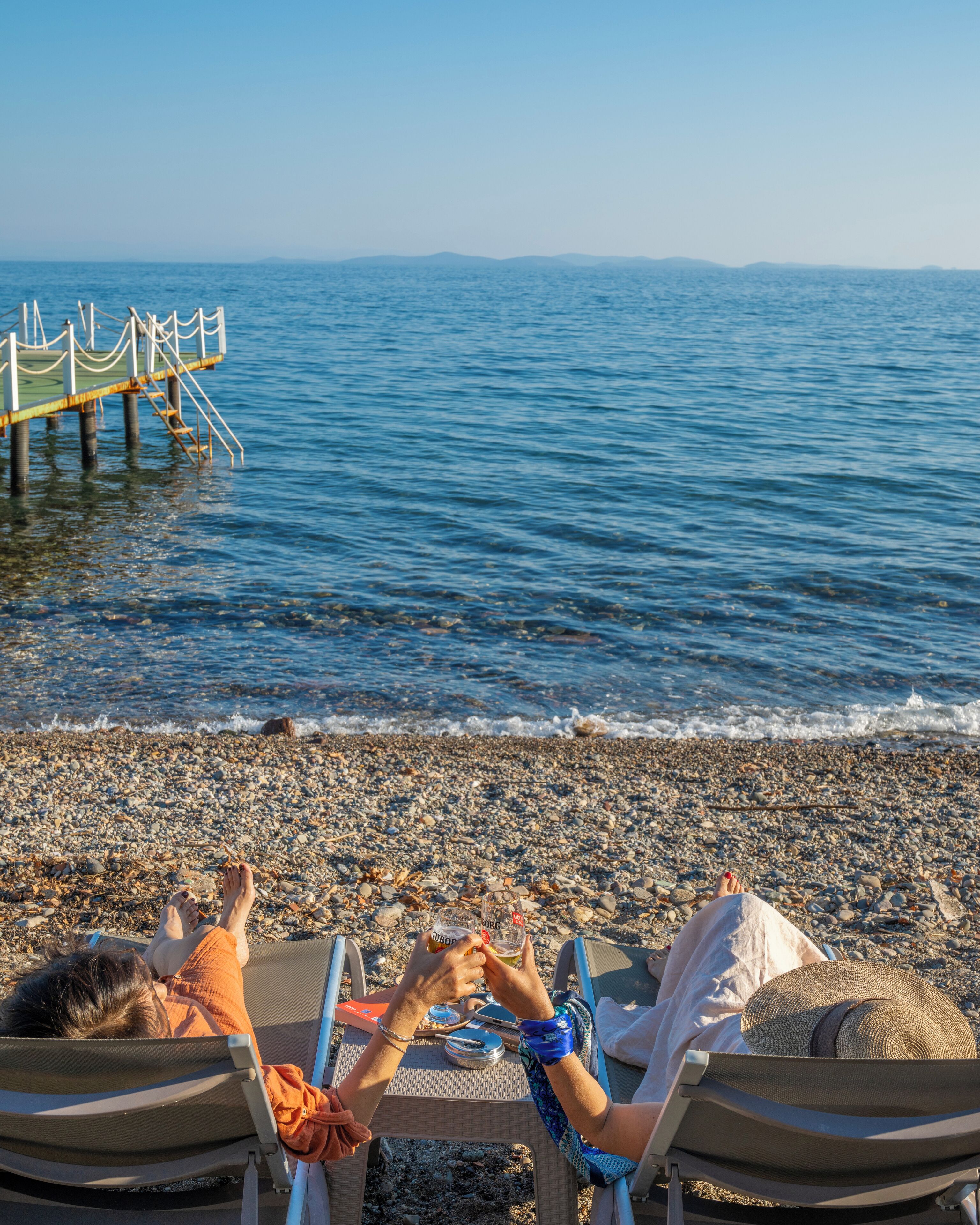 Een strand met een houten steiger die zich uitstrekt over de zee, en mensen die ontspannen in strandstoelen met uitzicht op het water.