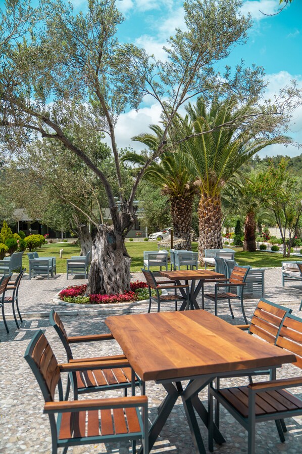 An outdoor seating area with wooden tables and chairs, surrounded by palm trees and other greenery. - Assos Kayalar Blue Beach Hotel (Ayvacik)