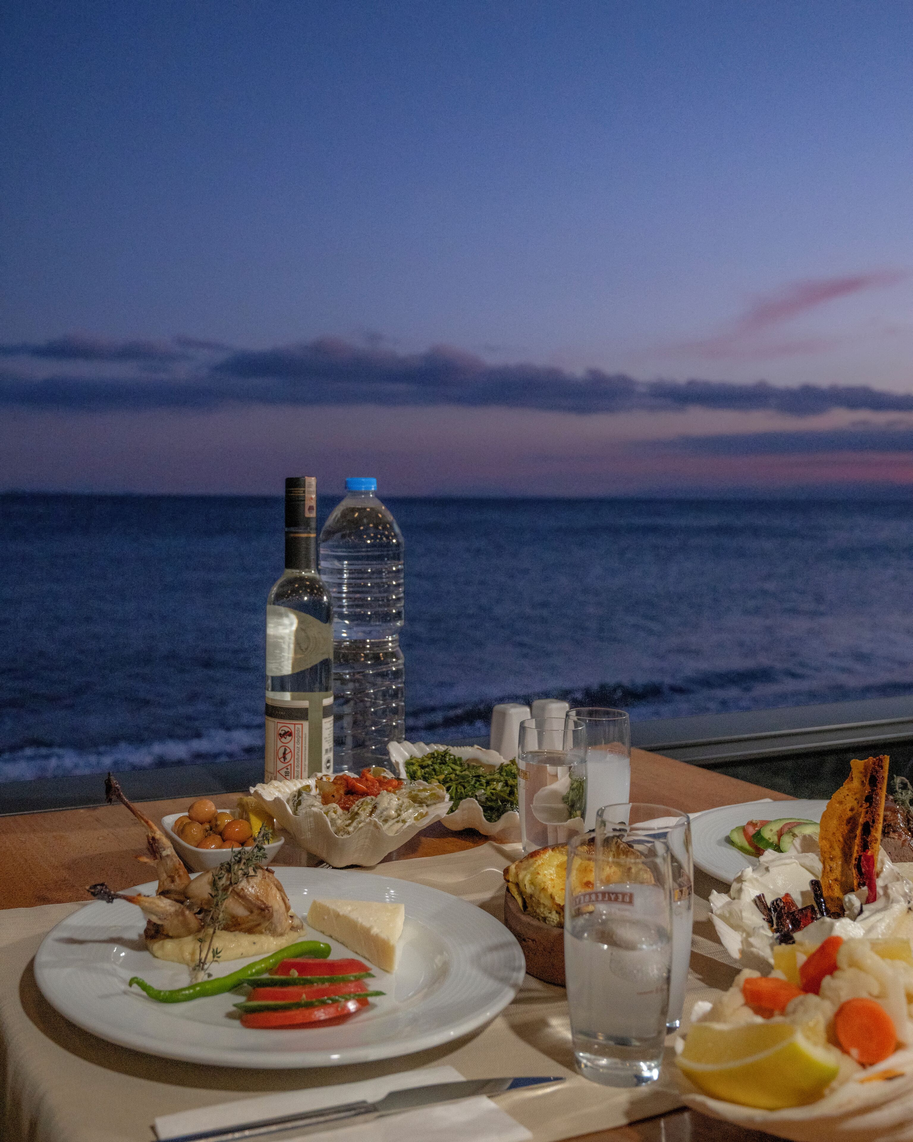 A table set with various dishes, including a roasted chicken leg, salad, and a dessert, overlooking a body of water at dusk.