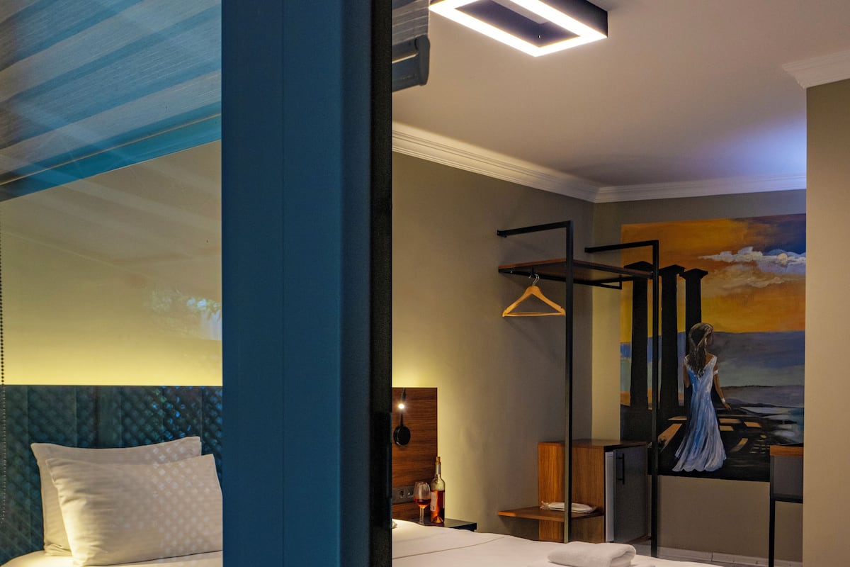 A hotel room with a large bed, a bedside table with a lamp, a hanging coat rack, and a painting of a sunset on the wall.