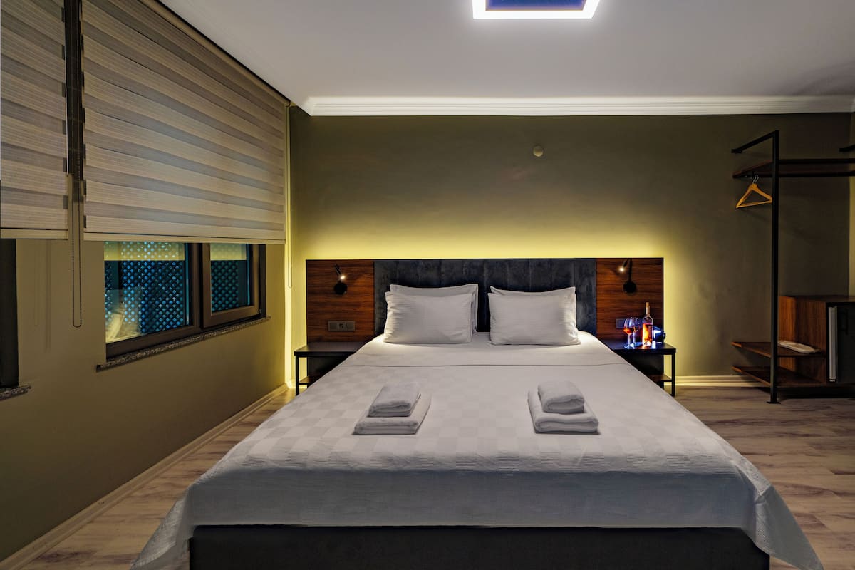 A double bed with two pillows, a bedside table with a bottle and glasses, a nightstand, a window with blinds, and a hanging light fixture.