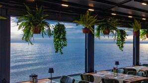 En restaurant med terrasse og havudsigt, med dækket bord, hængende planter og udsigt over vandet.