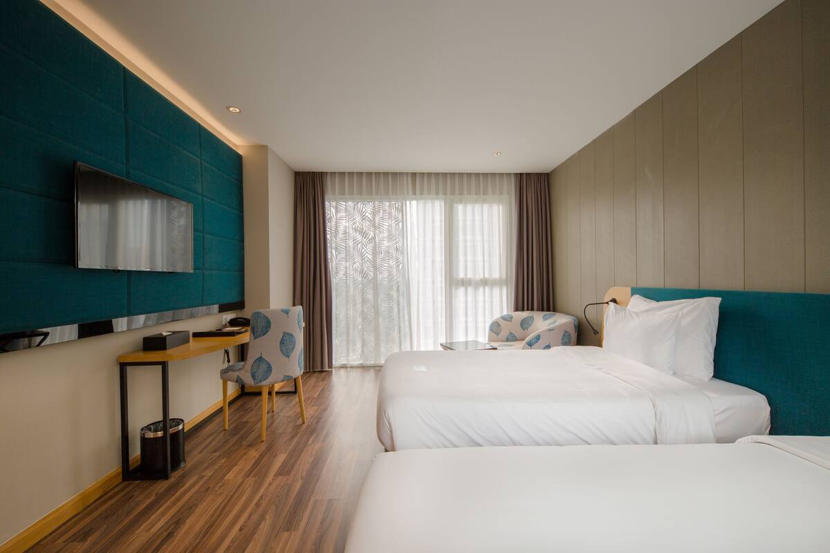 Quarto de hotel com duas camas, uma mesa, uma cadeira, uma televisão e uma janela grande com cortinas.