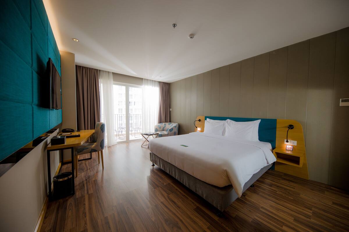 Quarto de hotel com uma cama grande, uma escrivaninha com cadeira, televisão na parede e uma janela com cortinas.