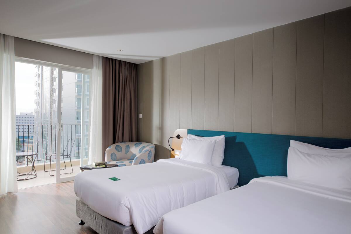 Quarto de hotel com duas camas, uma varanda com cadeira e vista para a cidade.