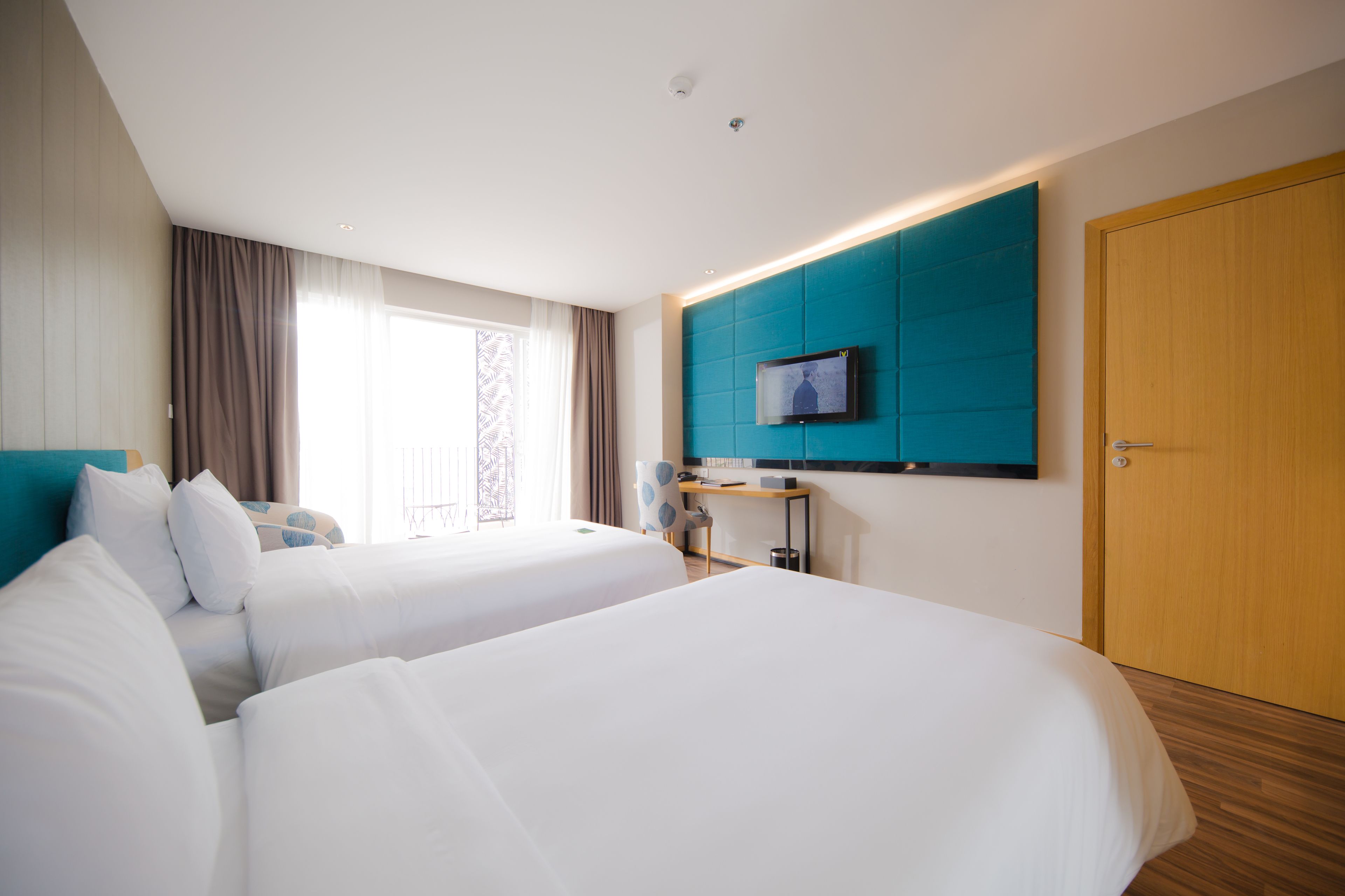 Habitación de hotel con dos camas, un televisor montado en una pared verde azulado, una puerta de madera y una ventana con cortinas.