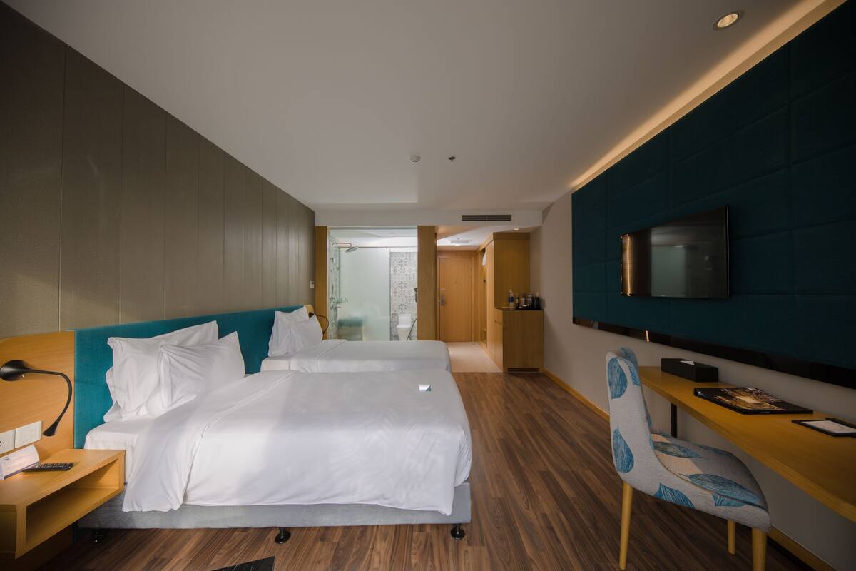 Quarto de hotel com uma cama grande, uma mesa, uma cadeira, uma televisão e um banheiro visível através de uma porta aberta.