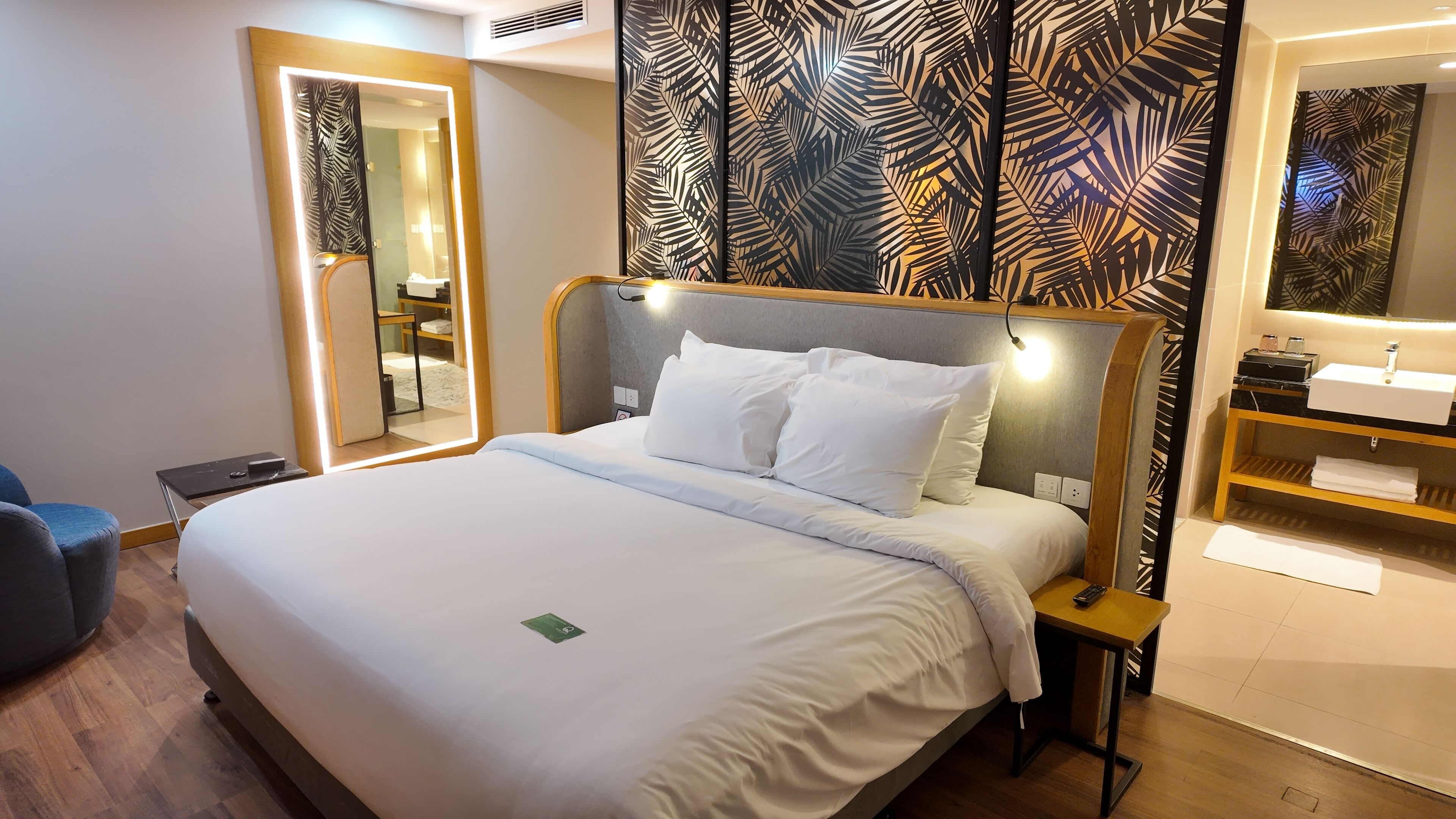 A hotel room with a large bed, a bedside table with a remote control, a mirror, and a bathroom with a sink and towel rack.